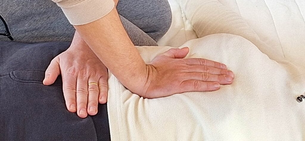 Primo piano delle mani di una persona, una delle quali indossa un anello d'oro, che esegue un massaggio sulla schiena di un'altra persona durante un Open Day alla scuola di Shiatsu. Entrambe le persone indossano abiti casual e di colore chiaro.