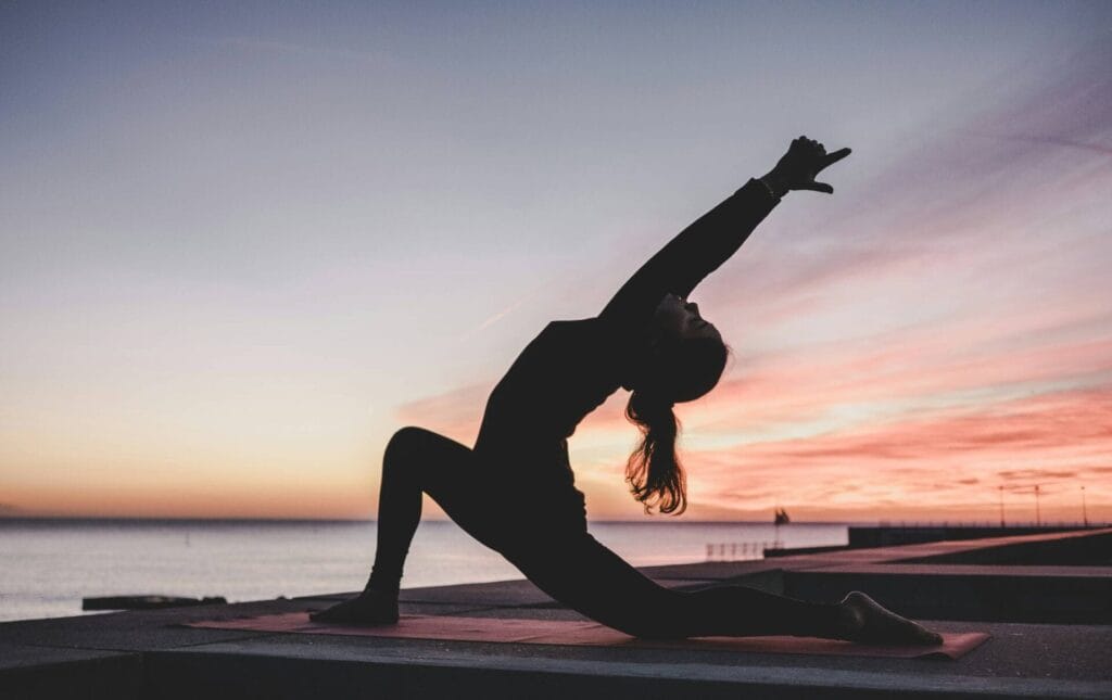 Una persona in posizione yoga al tramonto vicino a uno specchio d'acqua, con la sua silhouette in contrasto con il cielo colorato, si gode un momento di pace e serenità durante la sua lezione di prova gratuita.